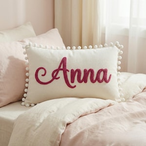 Puede incluir: Una almohada rectangular de color crema con el nombre "Anna" bordado en una fuente cursiva magenta. La almohada está adornada con pompones blancos. La almohada está sobre una cama con ropa de cama rosa.