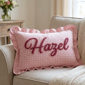 May include: A rectangular pink gingham pillow with the name "Hazel" embroidered in a script font. The pillow has a ruffled edge and is placed on a beige sofa. A cozy home decor item.