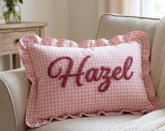 Può includere: Un cuscino rettangolare rosa a quadretti vichy con il nome "Hazel" ricamato in corsivo. Il cuscino ha un bordo a balze ed è posto su un divano beige. Un elemento di arredo per la casa.
