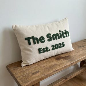 May include: Cream-colored rectangular pillow with the text "The Smith Est. 2025" in dark green, displayed on a rustic wooden bench. The pillow has a soft, textured appearance, and the lettering is raised.