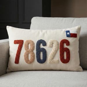 Könnte beinhalten: Cremefarbenes rechteckiges Kissen mit den in verschiedenen Farben gestickten Zahlen "78626". Eine kleine Texas-Flagge ist in der oberen rechten Ecke aufgenäht. Das Kissen liegt auf einem hellgrauen Sofa.