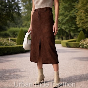 May include: A brown suede midi skirt with a front slit, paired with beige heeled boots. The skirt has a high waist and a classic design. A white handbag is held in the left hand. The text "Urban Hide Fashion" is at the bottom.