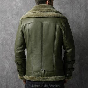 May include: Olive green leather jacket with a shearling collar and lining. The jacket has a button closure at the waist and buckle details on the sleeves. The back of the jacket features vertical stitching. The text "Urban Hide Fashion" is at the bottom.