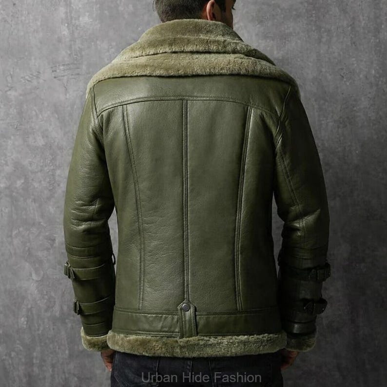 May include: Olive green leather jacket with a shearling collar and lining. The jacket has a button closure at the waist and buckle details on the sleeves. The back of the jacket features vertical stitching. The text "Urban Hide Fashion" is at the bottom.