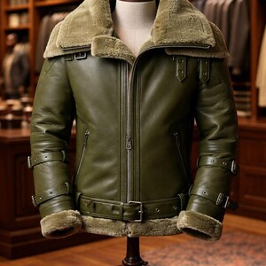 May include: An olive green leather jacket with a shearling collar and cuffs. The jacket has a zip closure, multiple buckles, and a waist belt. The jacket is displayed on a mannequin.