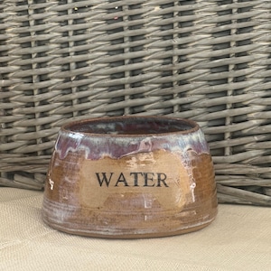May include: A round, tapered ceramic bowl with a wide base and a glazed finish in shades of brown and purple. The word WATER is printed in black, sans-serif capital letters on the front of the bowl. The interior and rim feature a glossy glaze.