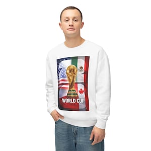 May include: White sweatshirt featuring a graphic of the World Cup trophy, with the flags of the United States, Mexico, and Canada. The words "WORLD CUP" are printed in white. The sweatshirt is worn by a person.