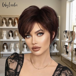 May include: A short, dark auburn wig with side-swept bangs. The wig is styled with a layered cut. The image includes the text "Wig Landia" in the upper left corner. Shelves of wigs are visible in the background.