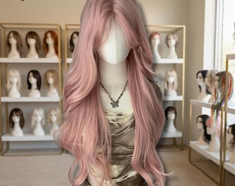 Puede incluir: Una peluca larga y ondulada de color rosa claro sobre un maniquí. La peluca tiene una raya a un lado y flequillo. El maniquí lleva un collar con un colgante de mariposa. El fondo muestra estanterías con otras pelucas y el texto "Wig Landia".