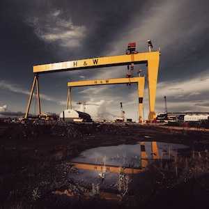 May include: Two large yellow gantry cranes stand prominently against a dark, cloudy sky. Each crane features the black text H & W on its horizontal beam. The industrial structures are positioned over a dark, uneven ground with a puddle reflecting them.