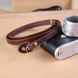 Puede incluir: Una correa de cámara de cuero marrón con un anillo de metal y un remache de latón. La correa está en bucle y se encuentra junto a una cámara negra y plateada. La correa está hecha de una sola pieza de cuero.