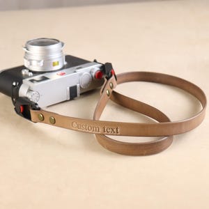 Peut inclure: Une sangle de caméra en cuir marron avec le texte "Custom text" en relief. La sangle est attachée à un appareil photo argenté et noir. La sangle a des rivets couleur laiton et une boucle noire.