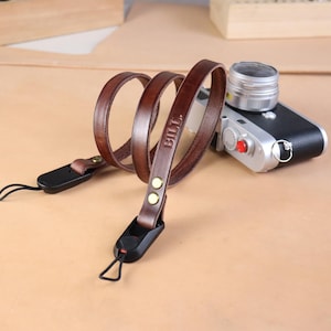 Op de afbeelding: Een bruinleren camerariem met de naam "BILL" erin geëmbosseerd. De riem is bevestigd aan een zwarte connector met een kleine zwarte lus. Op de achtergrond staat een zilveren en zwarte camera.