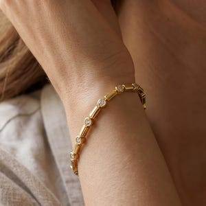 Puede incluir: Una pulsera de cadena dorada que presenta una serie de eslabones metálicos cilíndricos que se alternan con pequeñas piedras redondas y transparentes engastadas en biseles circulares. La pulsera tiene un acabado pulido y un patrón repetitivo.