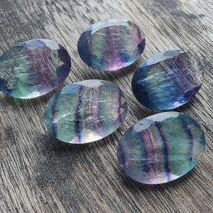 Può includere: Cinque gemme ovali sfaccettate con sfumature di blu, verde e viola. Sono disposte su una superficie di legno grigio invecchiato. Le pietre hanno una superficie lucida e riflettente, probabilmente utilizzate per la creazione di gioielli.