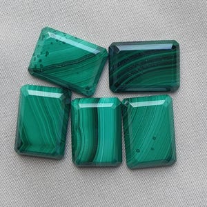 Puede incluir: Cinco piedras preciosas de malaquita rectangulares con superficies pulidas, que muestran distintos patrones de bandas en verde oscuro y claro. Las piedras están dispuestas en un grupo, cada una con bordes facetados y un acabado suave y brillante.