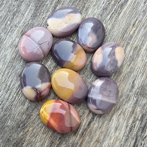 Puede incluir: Colección de piedras ovaladas pulidas en tonos morados, amarillos y marrones. Las piedras tienen un acabado brillante y patrones variados, dispuestos en forma circular. Las piedras están sobre una superficie de madera.