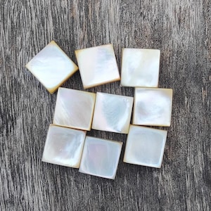 Könnte beinhalten: Neun quadratische Perlmuttfliesen mit schillernden Oberflächen und einem warmen, cremigen Farbton. Die Fliesen sind auf einer verwitterten, grauen Holzoberfläche angeordnet. Jede Fliese hat eine leicht abgerundete Kante.