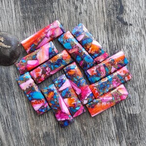 May include: A collection of rectangular gemstone beads in shades of pink, orange, blue, and gold. The beads are arranged on a wooden surface, showcasing their unique patterns and colors. These beads are ideal for jewelry making.