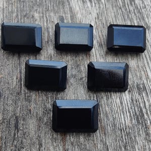 May include: Five rectangular, black gemstones with beveled edges are arranged on a weathered wooden surface. The gems have a polished, reflective surface, and their dark colour contrasts with the lighter wood grain.