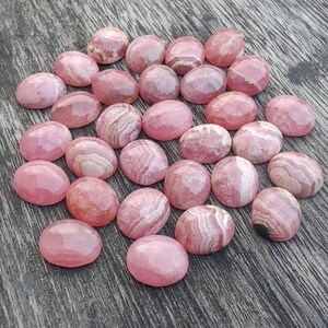 May include: A selection of pink and white gemstone cabochons. The oval-shaped stones have a polished, faceted surface. The pink hue varies, with some showing white banding. The gemstones are arranged on a wooden surface.