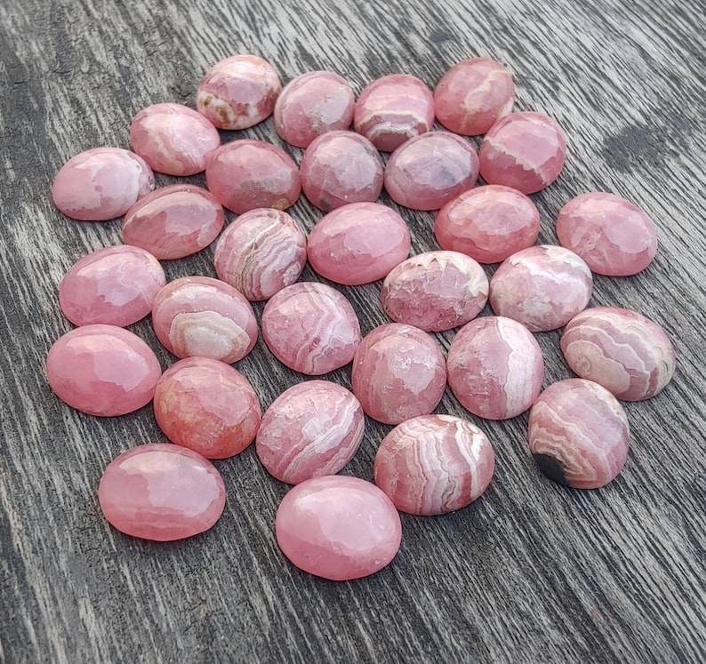 May include: A selection of pink and white gemstone cabochons. The oval-shaped stones have a polished, faceted surface. The pink hue varies, with some showing white banding. The gemstones are arranged on a wooden surface.