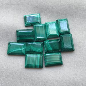 May include: A collection of ten rectangular malachite gemstones, each featuring distinct dark and light green banded patterns with a polished, faceted surface. The stones are arranged in a loose cluster, showcasing natural variations in the mineral.