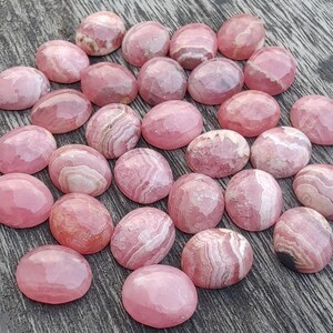 May include: A selection of pink rhodochrosite gemstones. The oval-shaped stones have a polished, faceted surface. The color ranges from light to deep pink, with white banding. The gemstones are arranged on a wooden surface.