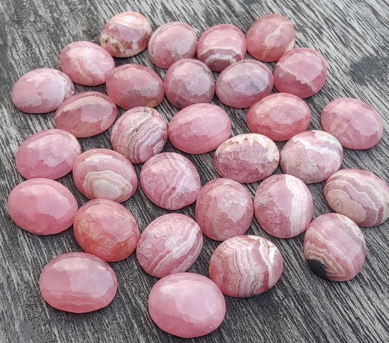 May include: A selection of pink rhodochrosite gemstones. The oval-shaped stones have a polished, faceted surface. The color ranges from light to deep pink, with white banding. The gemstones are arranged on a wooden surface.