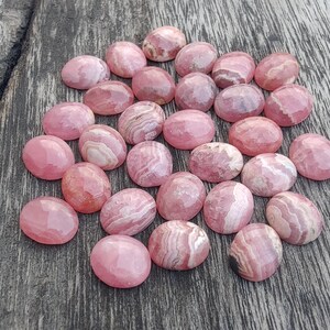 May include: A selection of pink rhodochrosite cabochons with a faceted surface. The stones range in colour from light to deep pink, with white and darker pink banding. The gemstones are oval-shaped and arranged on a wooden surface.