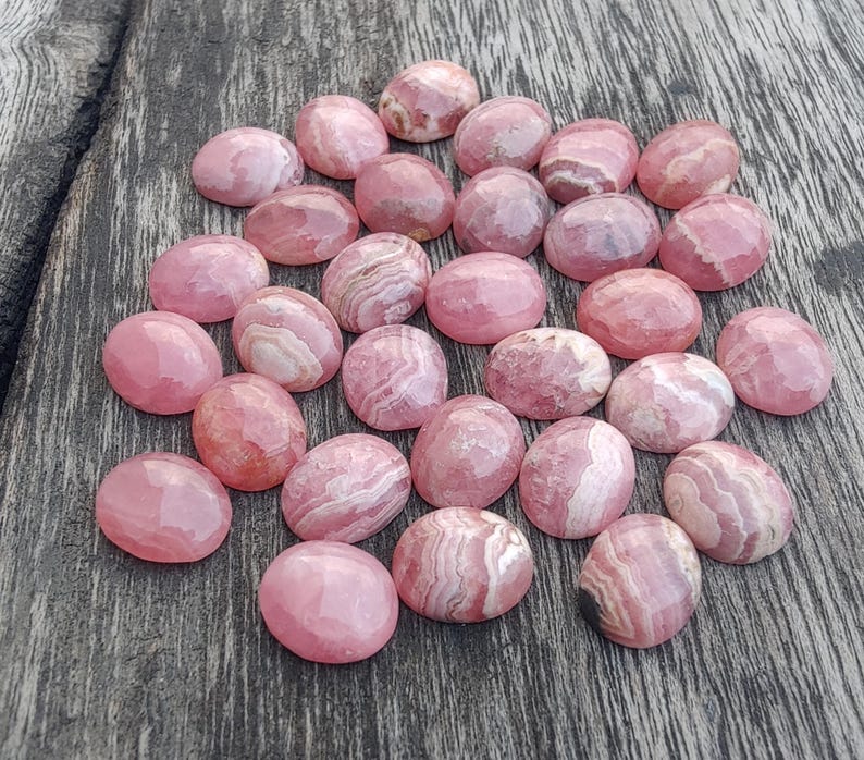 May include: A selection of pink rhodochrosite cabochons with a faceted surface. The stones range in colour from light to deep pink, with white and darker pink banding. The gemstones are oval-shaped and arranged on a wooden surface.