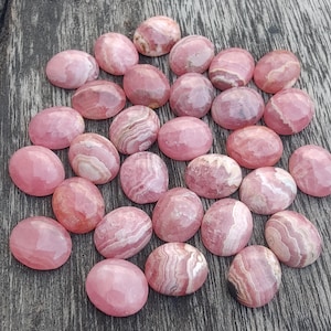May include: A selection of pink rhodochrosite cabochons, oval in shape, displaying diverse patterns of light and dark pink hues. The gemstones are presented on a wooden surface, highlighting their polished appearance and natural variations.