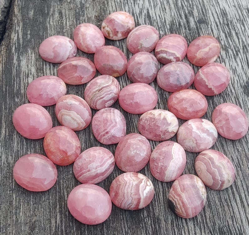 May include: A selection of pink rhodochrosite cabochons, oval in shape, displaying diverse patterns of light and dark pink hues. The gemstones are presented on a wooden surface, highlighting their polished appearance and natural variations.