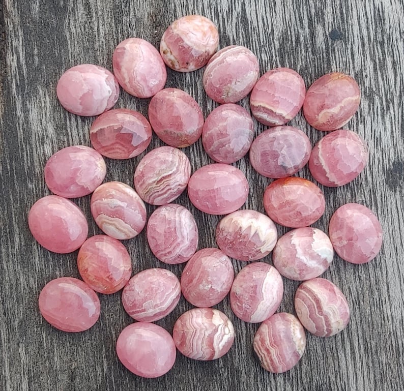 May include: A collection of oval-shaped, polished rhodochrosite gemstones. The stones display a range of pink hues, from pale to deep rose, with distinctive banding patterns. The gemstones are arranged on a weathered wooden surface.