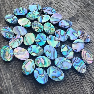 May include: A collection of oval-shaped abalone shell cabochons. The shells display iridescent colors of blue, green, and purple with unique swirling patterns. The cabochons are arranged on a dark wooden surface, showcasing their natural beauty.