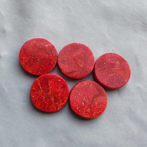 Pode incluir: Cinco cabochões de pedra vermelha, circulares e planos, com uma textura natural, porosa e manchada. Cada peça apresenta um tom carmesim vibrante com variações subtis na intensidade da cor e padrões orgânicos nas superfícies.