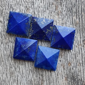 May include: Five square lapis lazuli gemstones with faceted pyramid tops. The deep blue stones have flecks of gold and are arranged on a wooden surface.