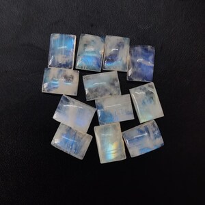 Könnte beinhalten: Eine Sammlung rechteckiger Mondstein-Cabochons. Die Steine haben eine durchscheinende, milchig-weiße Basis mit irisierenden blauen und gelben Blitzen. Die Steine sind auf einer dunklen Oberfläche angeordnet.