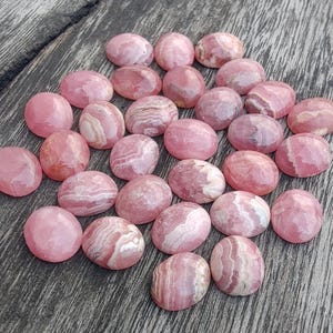 May include: A collection of pink and white oval gemstones with diverse patterns. The stones are arranged on a weathered wooden surface, highlighting their natural colours and textures. The gemstones have a polished appearance.