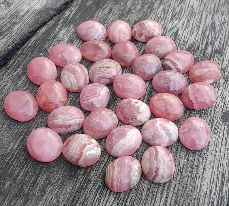 May include: A collection of pink and white oval gemstones with diverse patterns. The stones are arranged on a weathered wooden surface, highlighting their natural colours and textures. The gemstones have a polished appearance.