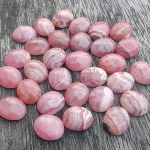 May include: A selection of pink cabochon gemstones with diverse patterns and hues. The oval-shaped stones are polished, displaying a smooth surface. The gemstones are arranged on a weathered wooden surface.