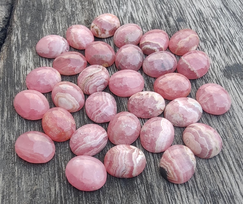 May include: A selection of pink cabochon gemstones with diverse patterns and hues. The oval-shaped stones are polished, displaying a smooth surface. The gemstones are arranged on a weathered wooden surface.