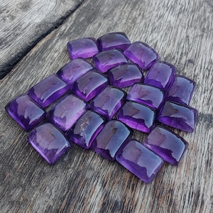 May include: A collection of polished, rectangular amethyst gemstones. The gems are a deep, rich purple color and have a smooth, reflective surface. They are arranged on a weathered wooden surface, showcasing their natural beauty.