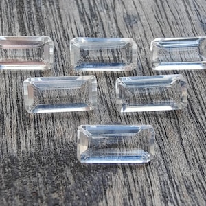 Puede incluir: Seis gemas rectangulares transparentes con bordes facetados dispuestas sobre una superficie de madera gris envejecida. Las gemas son transparentes, reflejan la luz y parecen estar cortadas con precisión. La estética general es de elegancia y claridad.
