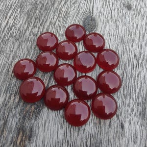 Könnte beinhalten: Eine Ansammlung von fünfzehn polierten, runden Cabochon-Edelsteinen in einem tiefroten Farbton. Die Steine sind auf einer verwitterten, grauen Holzoberfläche angeordnet, die ihre glatten, reflektierenden Oberflächen hervorhebt. Diese Cabochons eignen sich ideal für Schmuckherstellung oder Bastelprojekte.