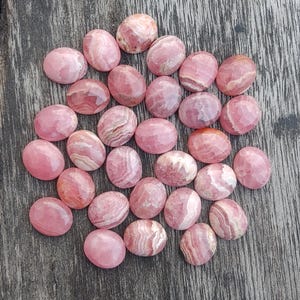 May include: A collection of oval-shaped, polished rhodochrosite gemstones. The stones display varying shades of pink and white, with some showing banded patterns. The gemstones are arranged on a weathered wooden surface.