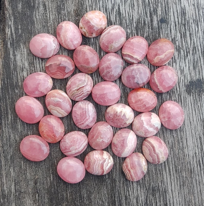 May include: A collection of oval-shaped, polished rhodochrosite gemstones. The stones display varying shades of pink and white, with some showing banded patterns. The gemstones are arranged on a weathered wooden surface.