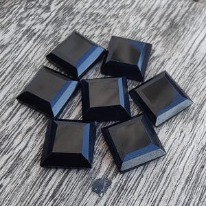 Puede incluir: Siete gemas negras de corte cuadrado dispuestas sobre una superficie de madera. Cada gema tiene una superficie pulida y reflectante y un borde biselado. Las gemas son probablemente para la fabricación de joyas u otras manualidades.