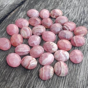 May include: A selection of pink and white cabochon gemstones, each with a rounded, oval shape. The stones are arranged on a weathered wooden surface, highlighting their natural patterns and colours. The gemstones vary in size and have a polished finish.