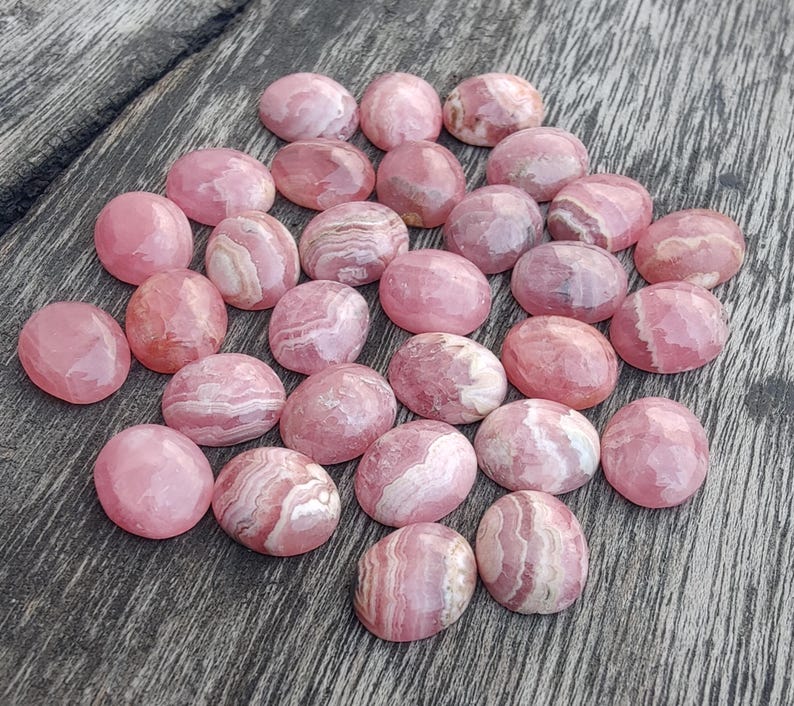 May include: A selection of pink and white cabochon gemstones, each with a rounded, oval shape. The stones are arranged on a weathered wooden surface, highlighting their natural patterns and colours. The gemstones vary in size and have a polished finish.